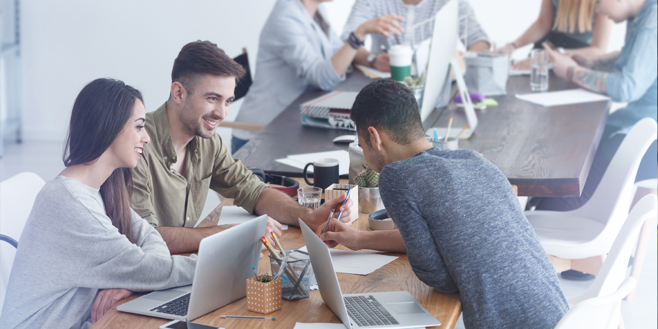 Three young interns sit at a table with notebooks and laptops working on their projects